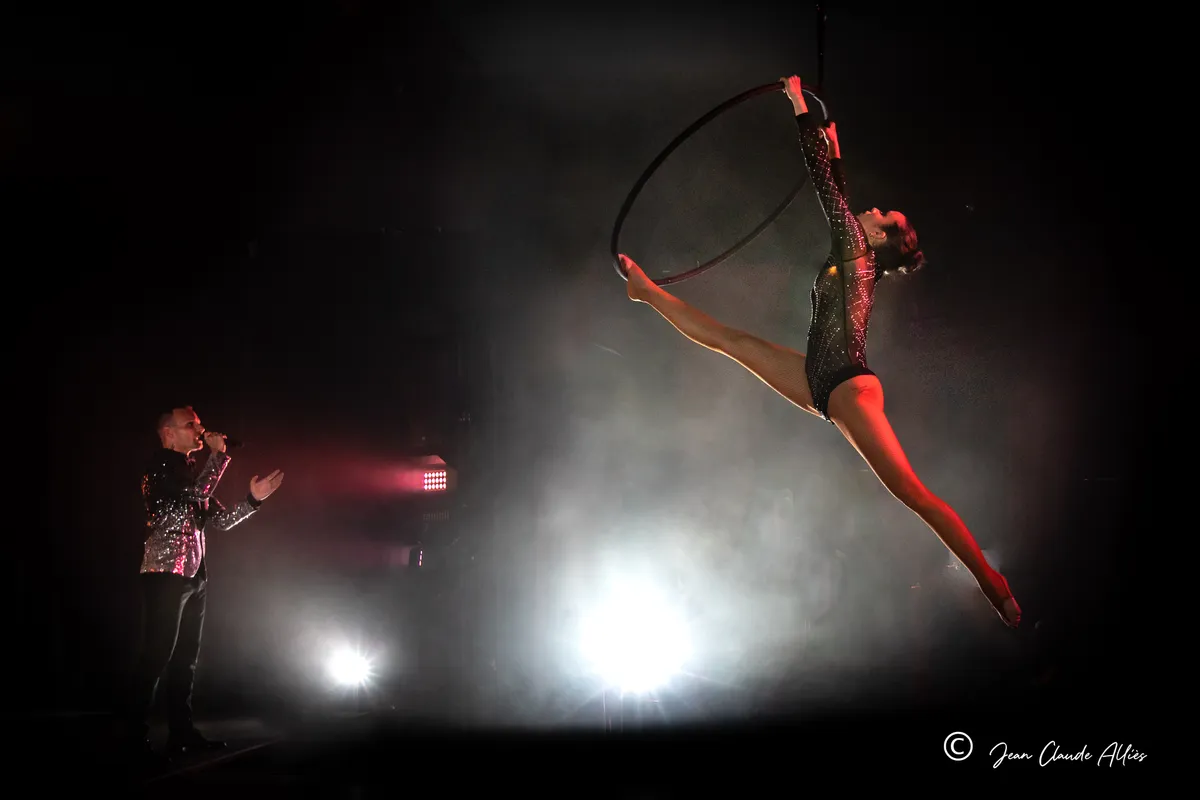 Acrobate en justaucorps noir étirant ses jambes depuis un cerceau aérien dans un contre-jour blanc.