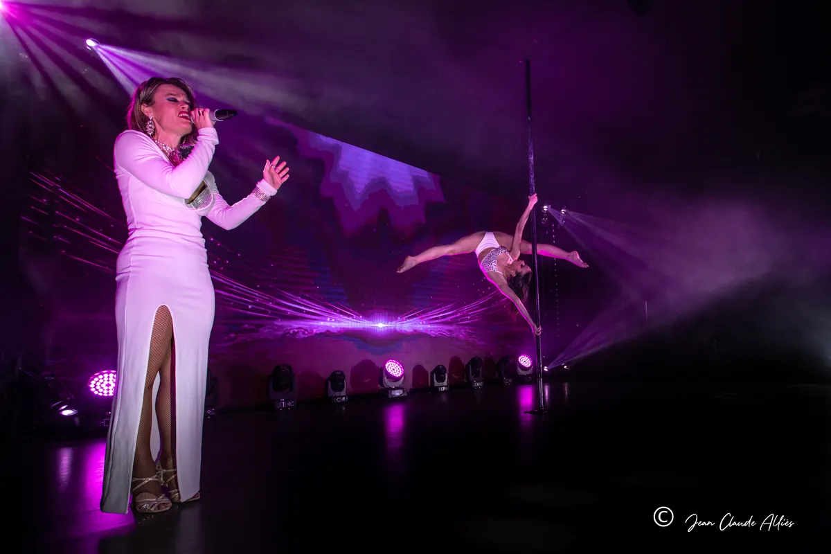 Chanteuse en longue robe blanche au premier plan avec une acrobate sur une barre dans une lumière violette.