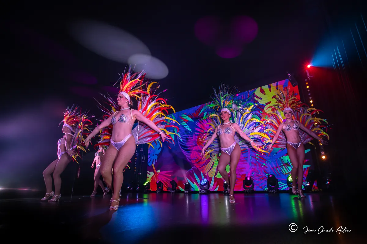 Danseuses aux grandes parures de plumes roses, jaunes et orange avançant devant un écran multicolore.