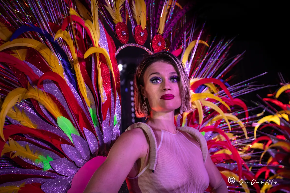 Portrait d'une danseuse encadrée d'une grande coiffe et de plumes roses, jaunes et orange.