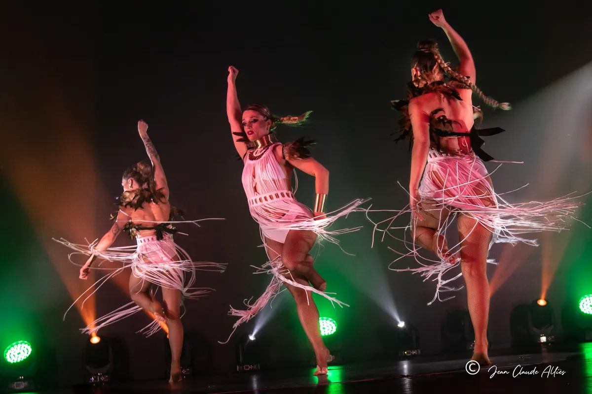 Trois danseuses levant un bras tandis que les longues franges claires de leurs costumes tourbillonnent.