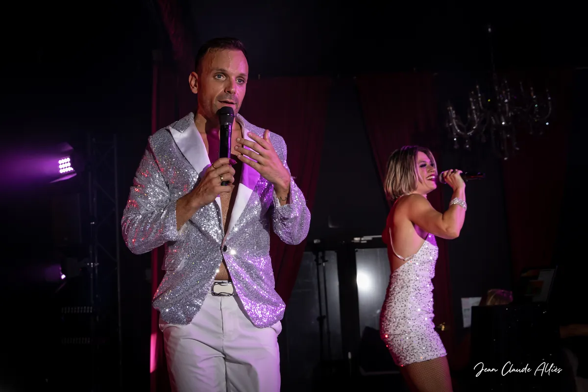 Chanteur en veste argentée au premier plan et chanteuse en robe brillante à ses côtés.