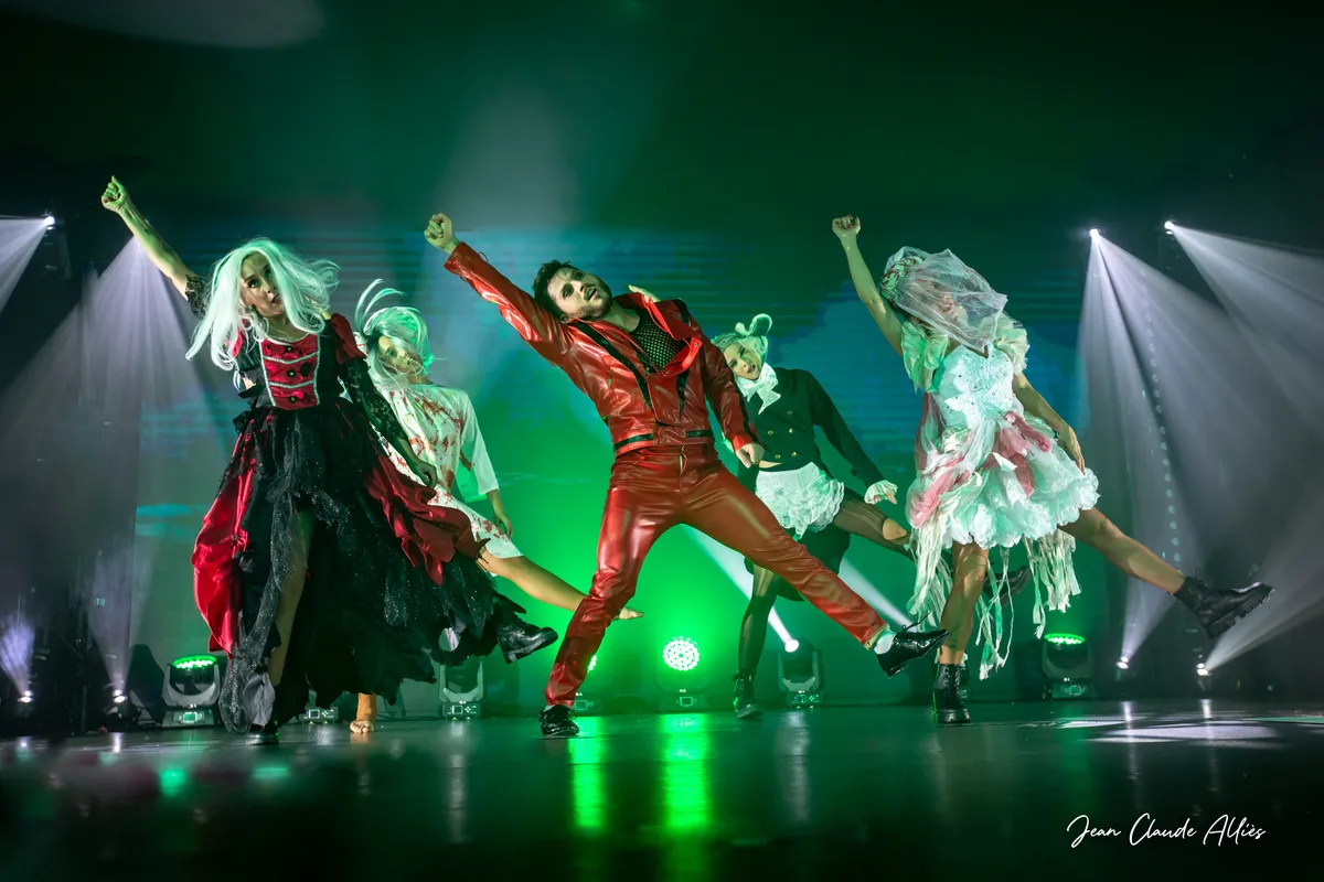 Danseur en costume rouge et danseuses aux perruques claires levant les bras sous une lumière verte.