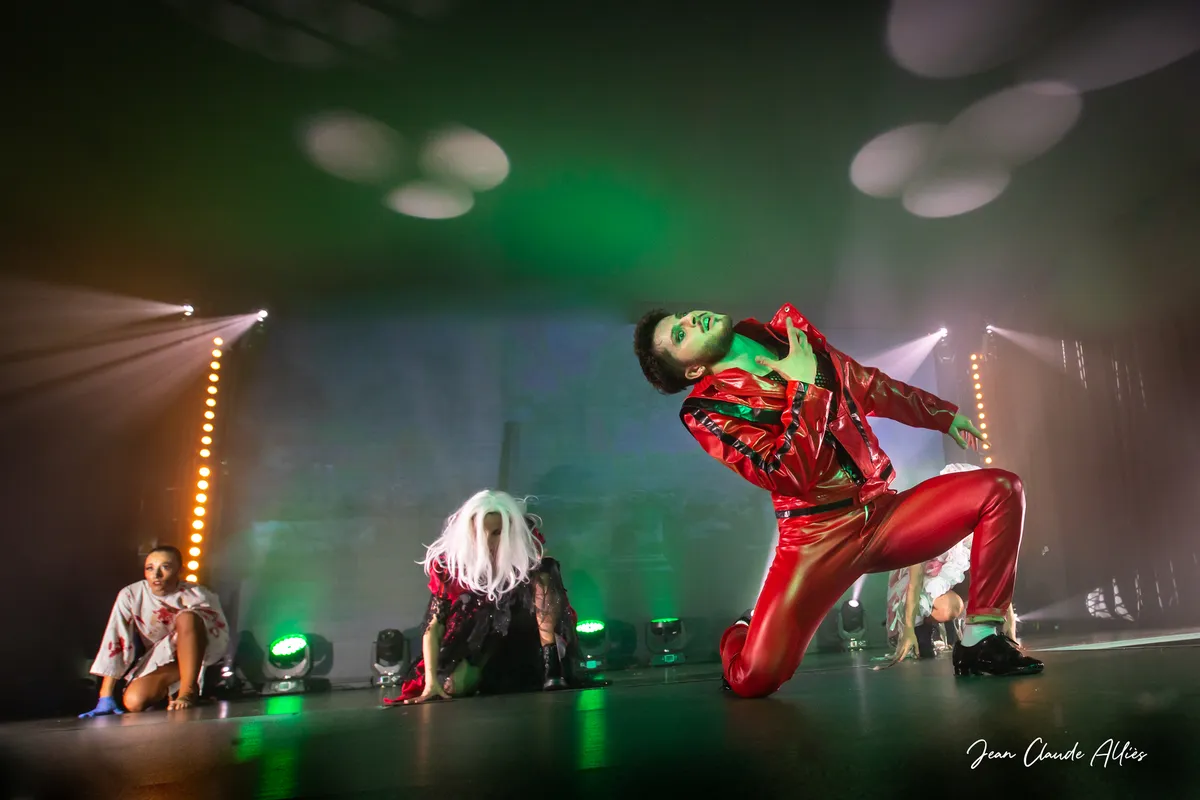 Danseur en costume rouge à genou, le buste incliné, devant des danseuses accroupies sur scène.
