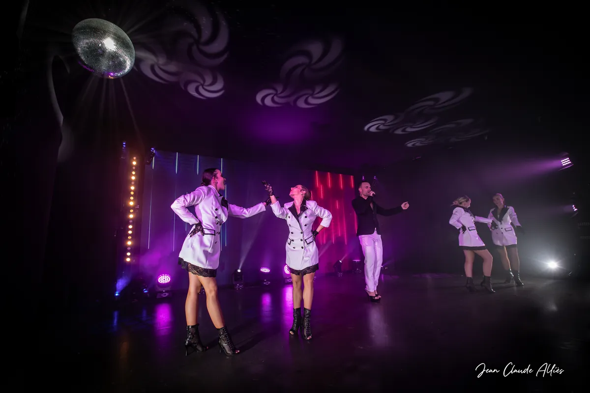 Troupe en vestes claires et shorts noirs sur une scène violette éclairée par une boule à facettes.