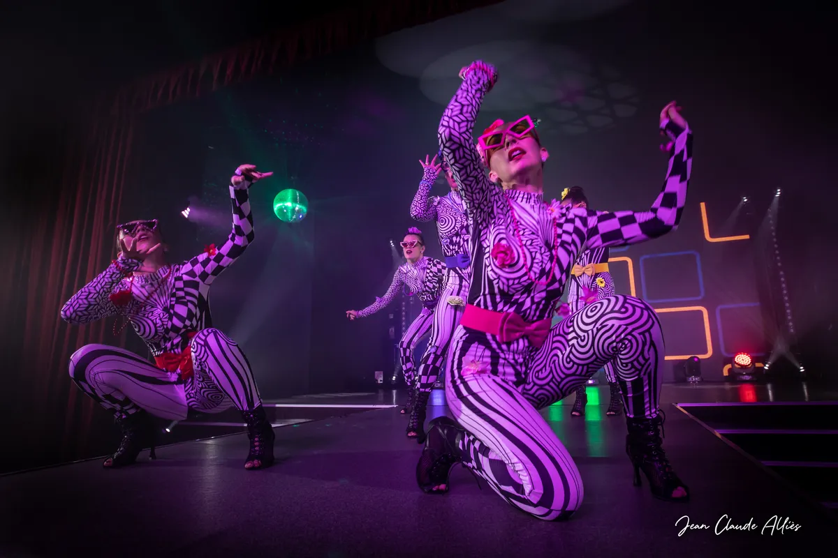 Danseuses accroupies en costumes graphiques noirs et blancs avec des lunettes roses et des ceintures colorées.