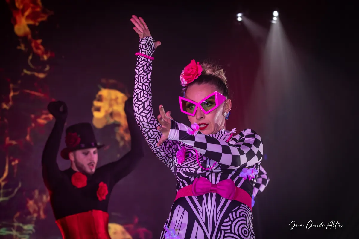 Danseuse aux lunettes roses levant un bras, vêtue de motifs noirs et blancs devant un danseur au chapeau noir.