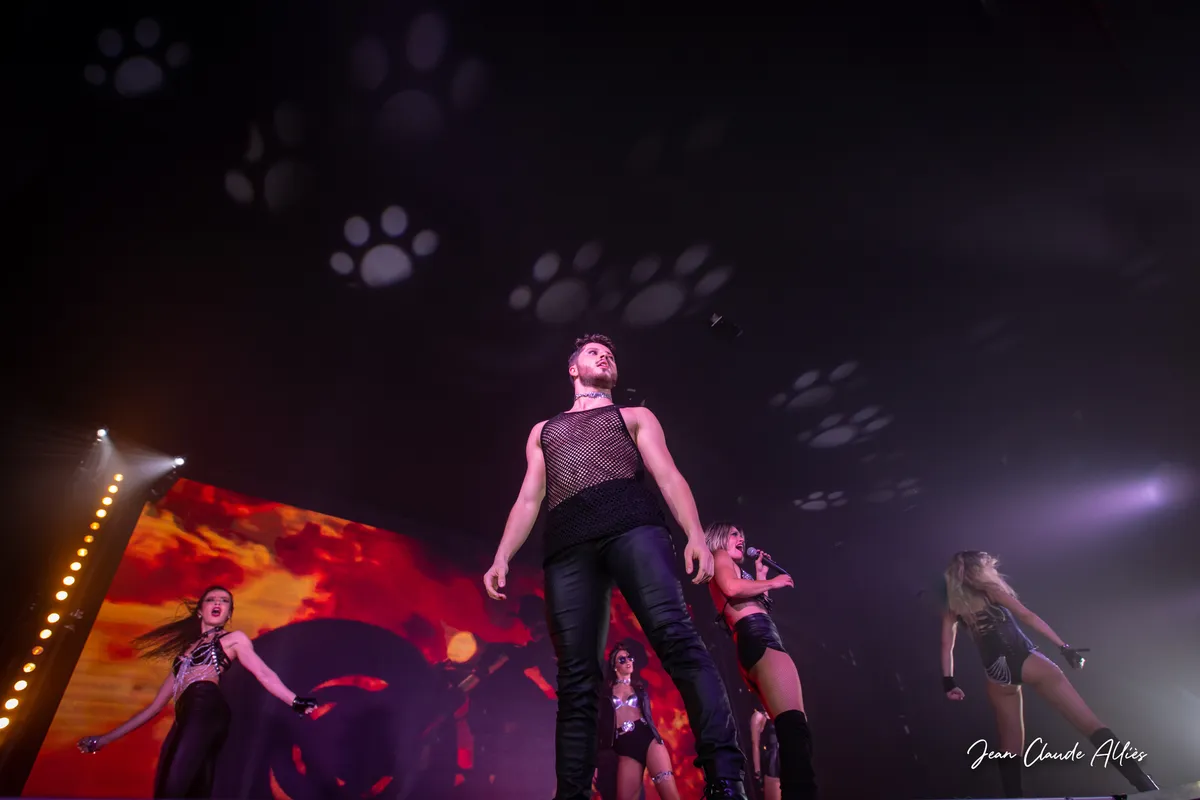 Danseur en débardeur noir cadré en contre-plongée, devant des danseuses et une projection de flammes.
