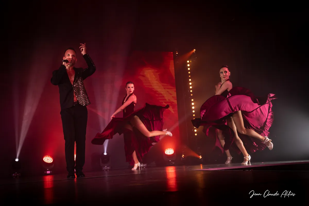 Chanteur en noir au micro devant deux danseuses qui soulèvent les volants de leurs jupes bordeaux.