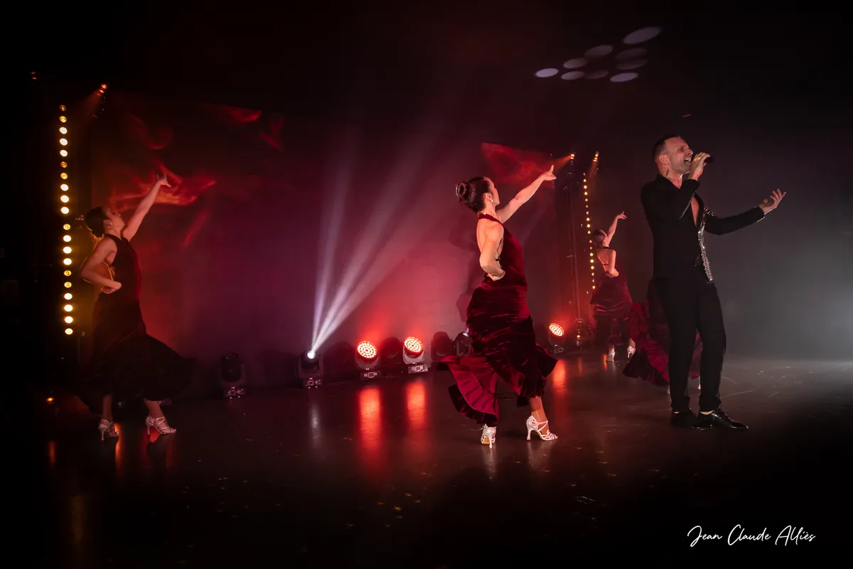 Chanteur et danseuses en longues jupes bordeaux, les bras déployés dans les faisceaux blancs.