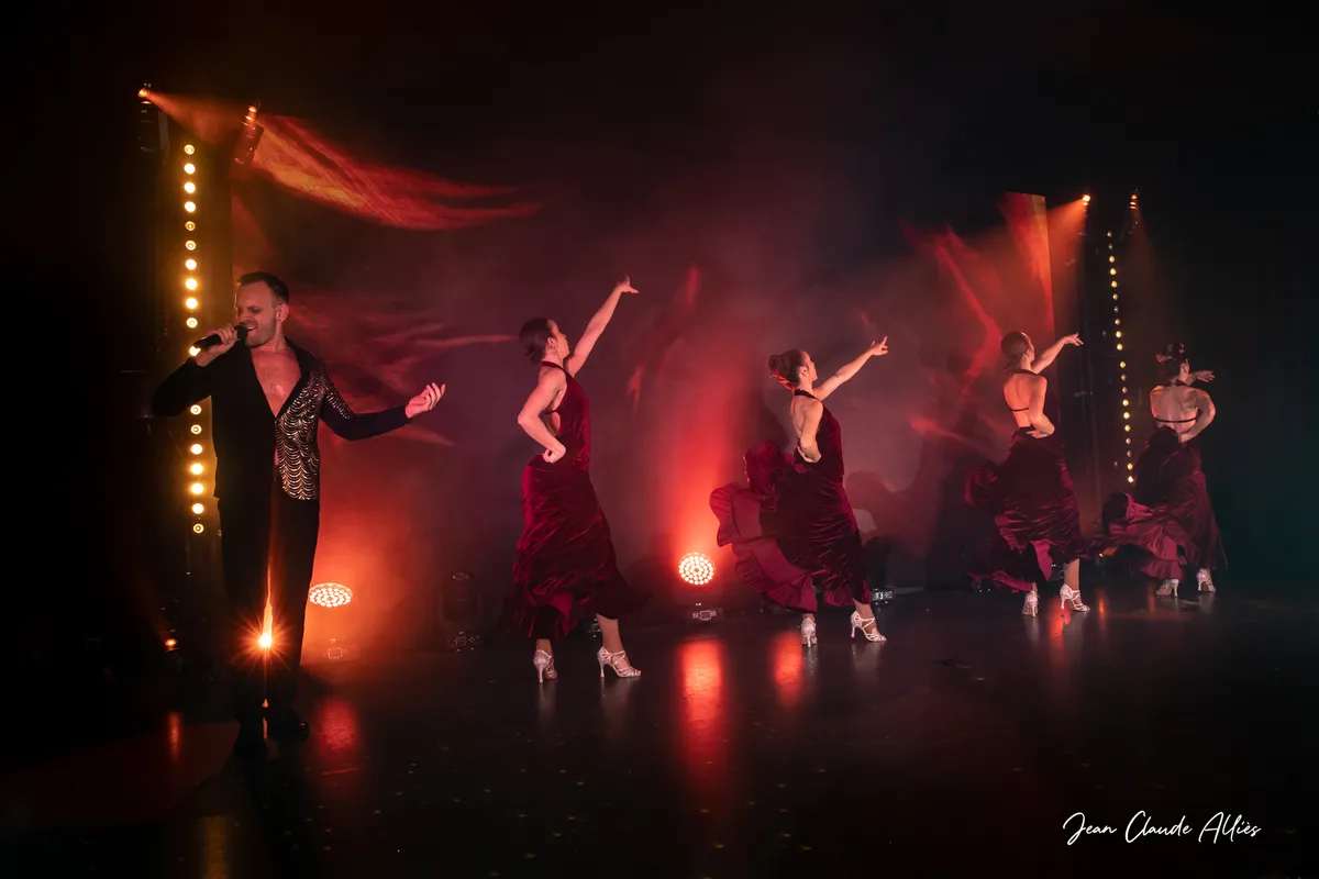 Chanteur au bord de la scène devant une ligne de danseuses en robes bordeaux, chacune levant un bras.
