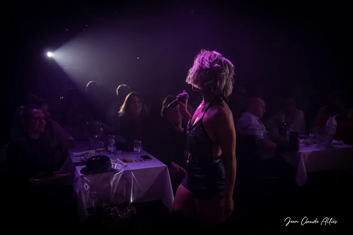 Chanteuse au micro près des tables du public, éclairée de profil par un projecteur violet.