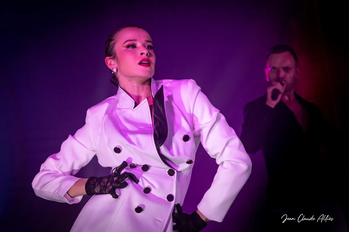 Danseuse en veste blanche croisée et gants noirs, une main à la taille sous une lumière rose.
