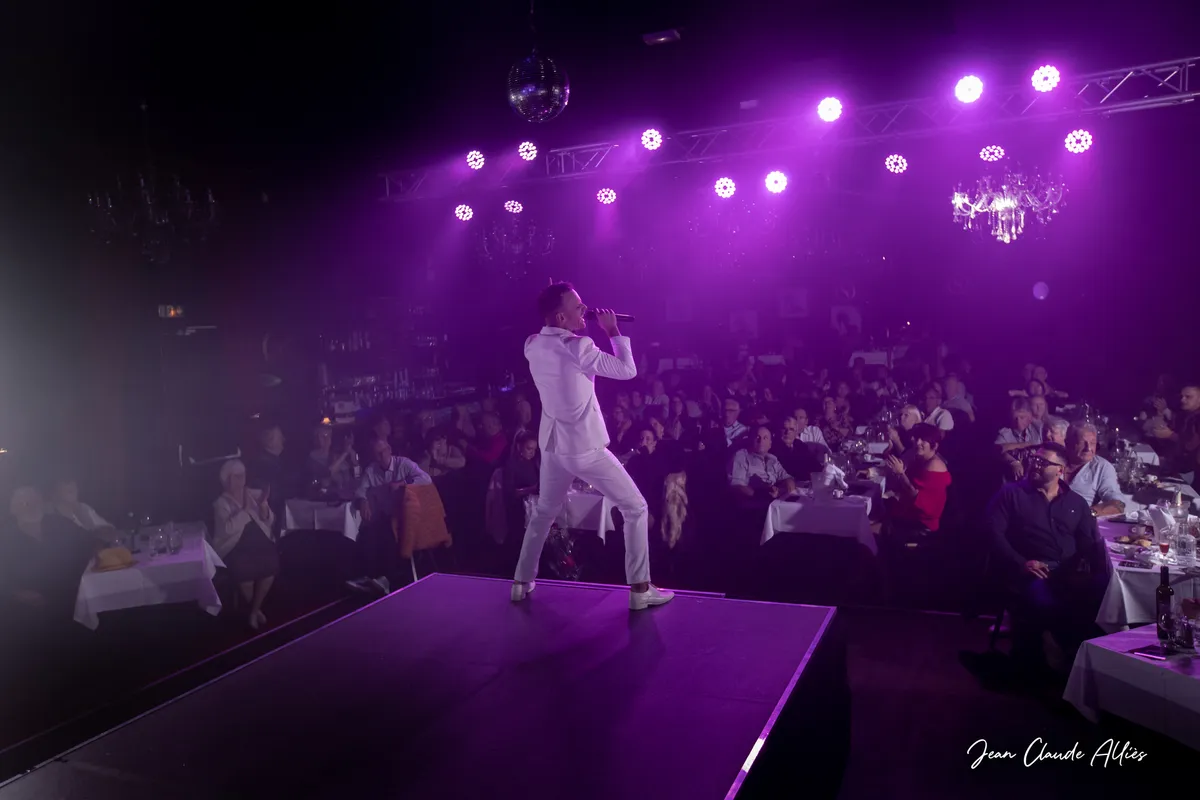 Chanteur en costume blanc vu de dos sur une avancée de scène au milieu du public attablé.