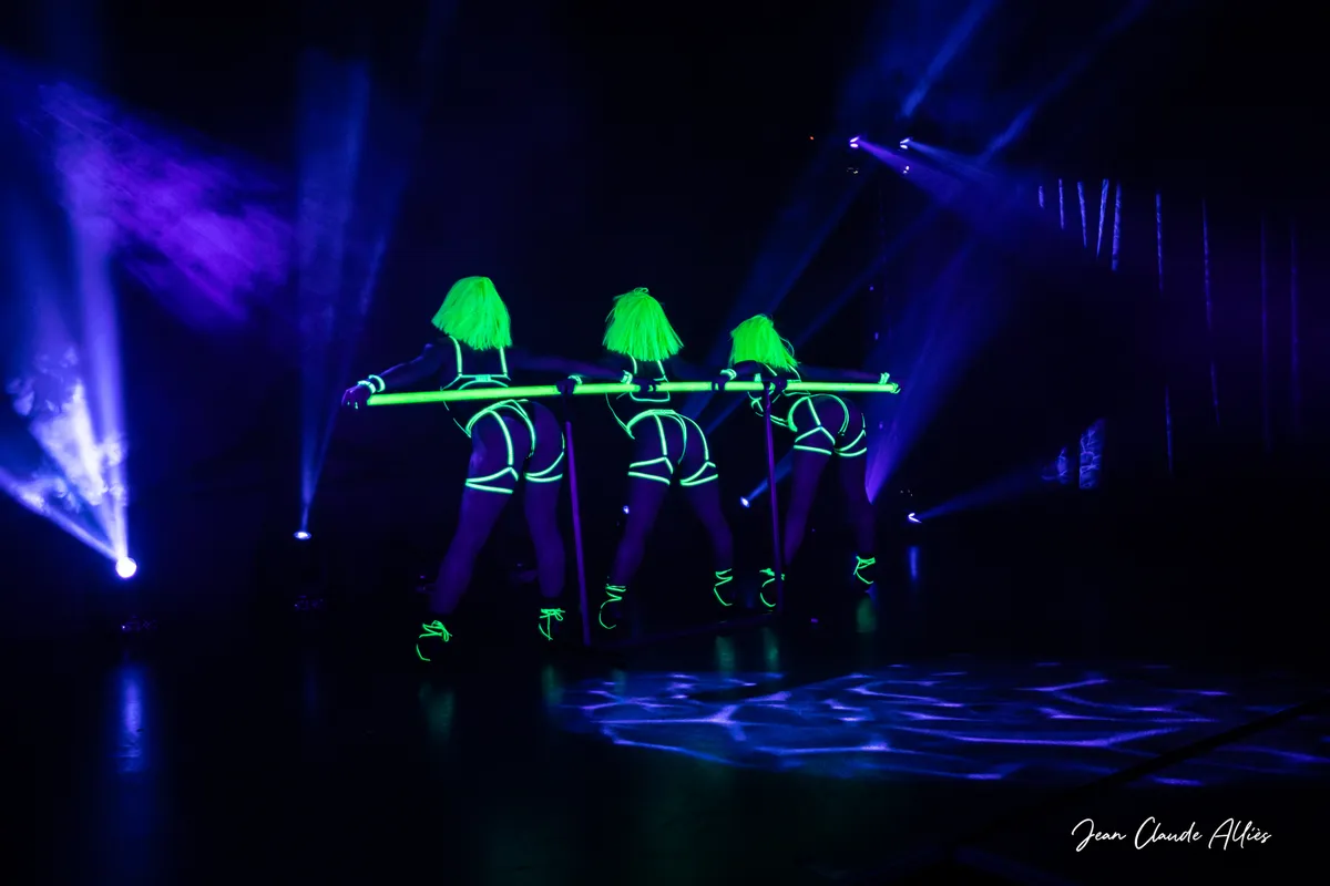 Trois danseuses aux perruques vertes et aux costumes fluorescents derrière une barre lumineuse sous éclairage bleu.