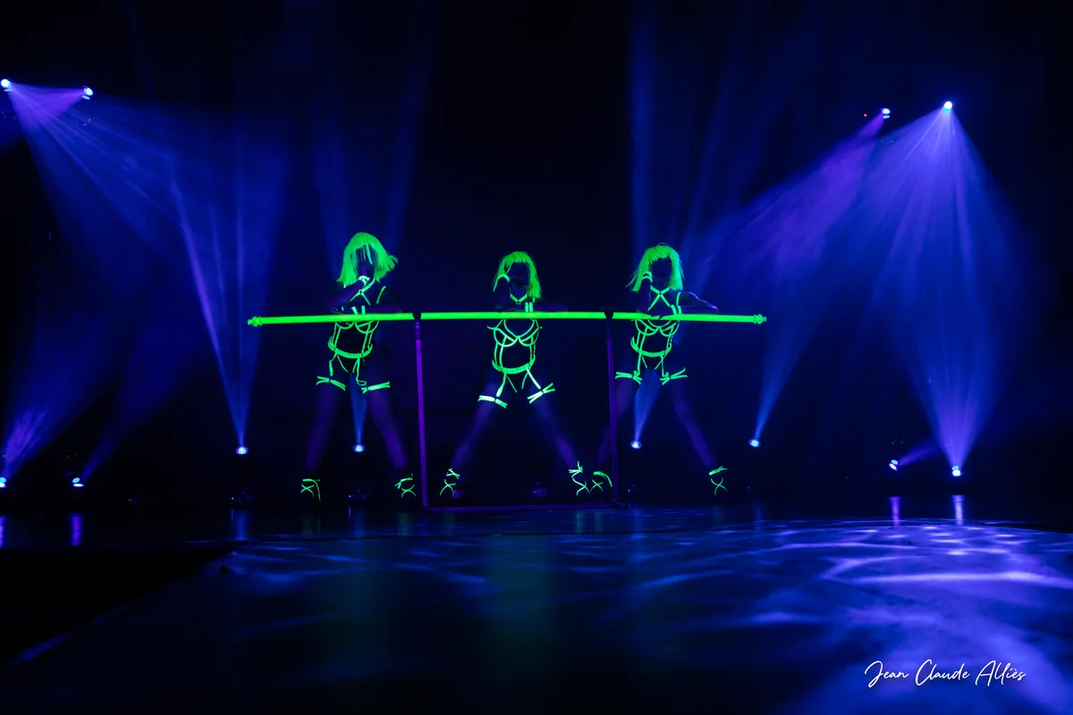 Vue d'ensemble de trois danseuses aux perruques vertes alignées derrière une barre fluorescente sur une scène bleue.