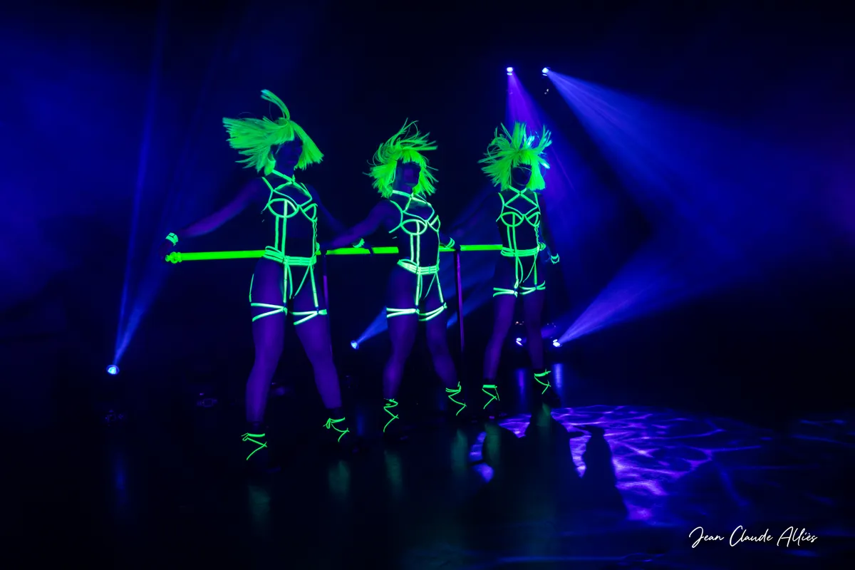 Trois danseuses faisant voler leurs perruques vertes derrière une barre fluorescente sur un fond bleu sombre.