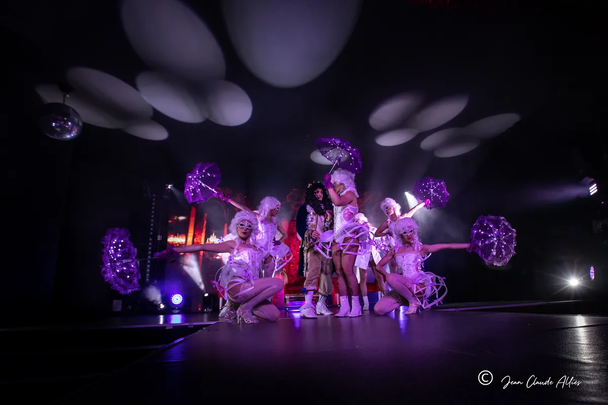 Danseuses en costumes argentés à paniers et perruques blanches autour d'un trône rouge, avec des ombrelles violettes.