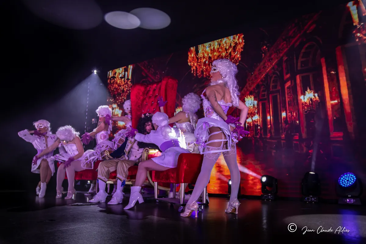 Troupe en perruques blanches et costumes de cour argentés réunie autour de grands fauteuils rouges.