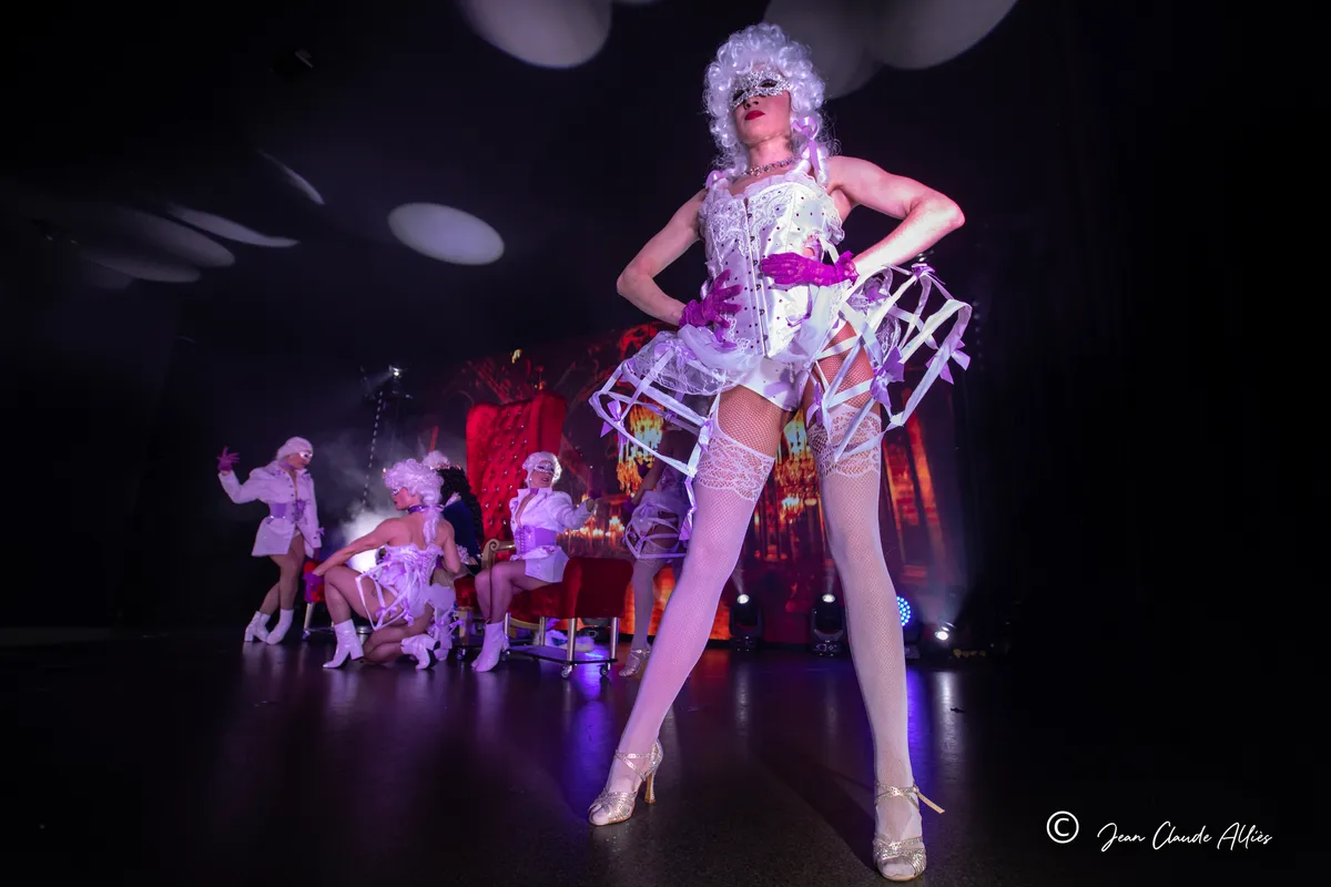 Danseuse en costume argenté à paniers, mains à la taille, devant la troupe en perruques blanches.