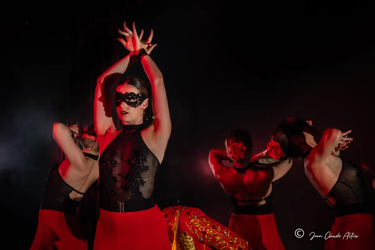 Danseuse portant un masque noir, les mains croisées au-dessus de la tête, devant des partenaires en rouge et noir.