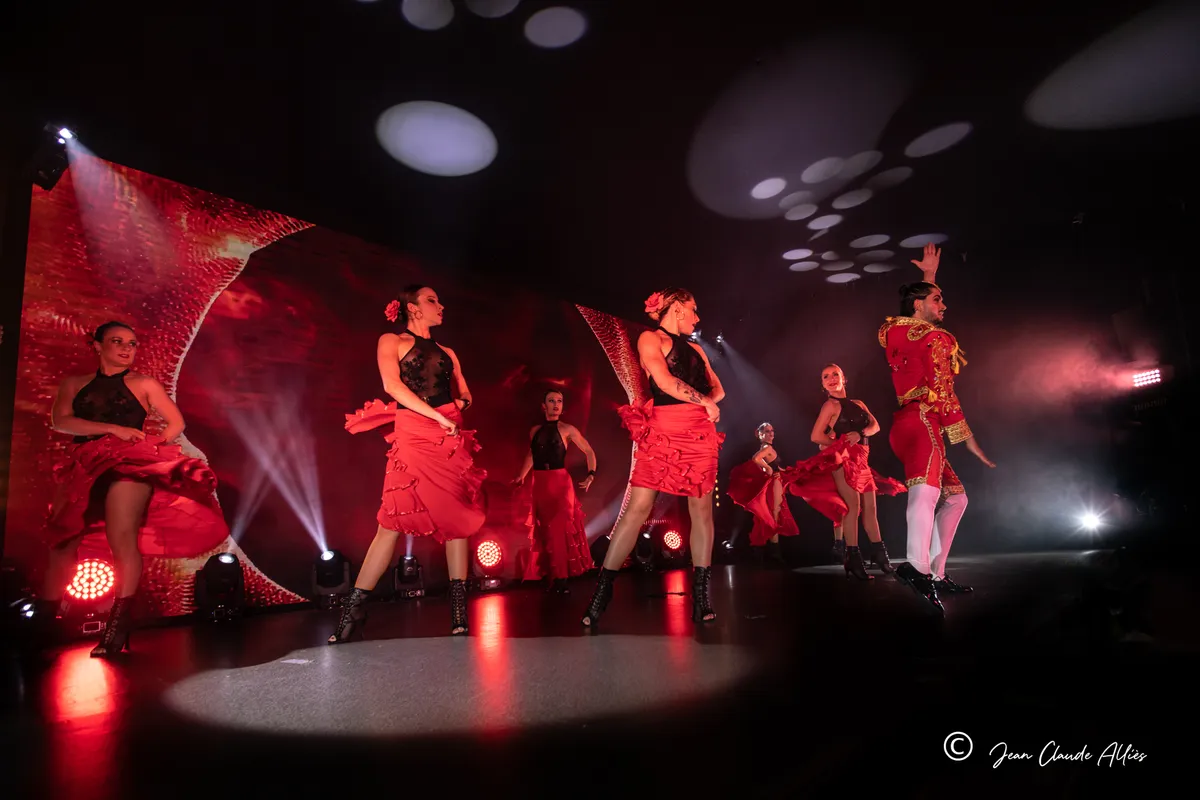 Chanteuse et troupe en costumes rouges dans un tableau d'ensemble éclairé par des faisceaux rouges et blancs.