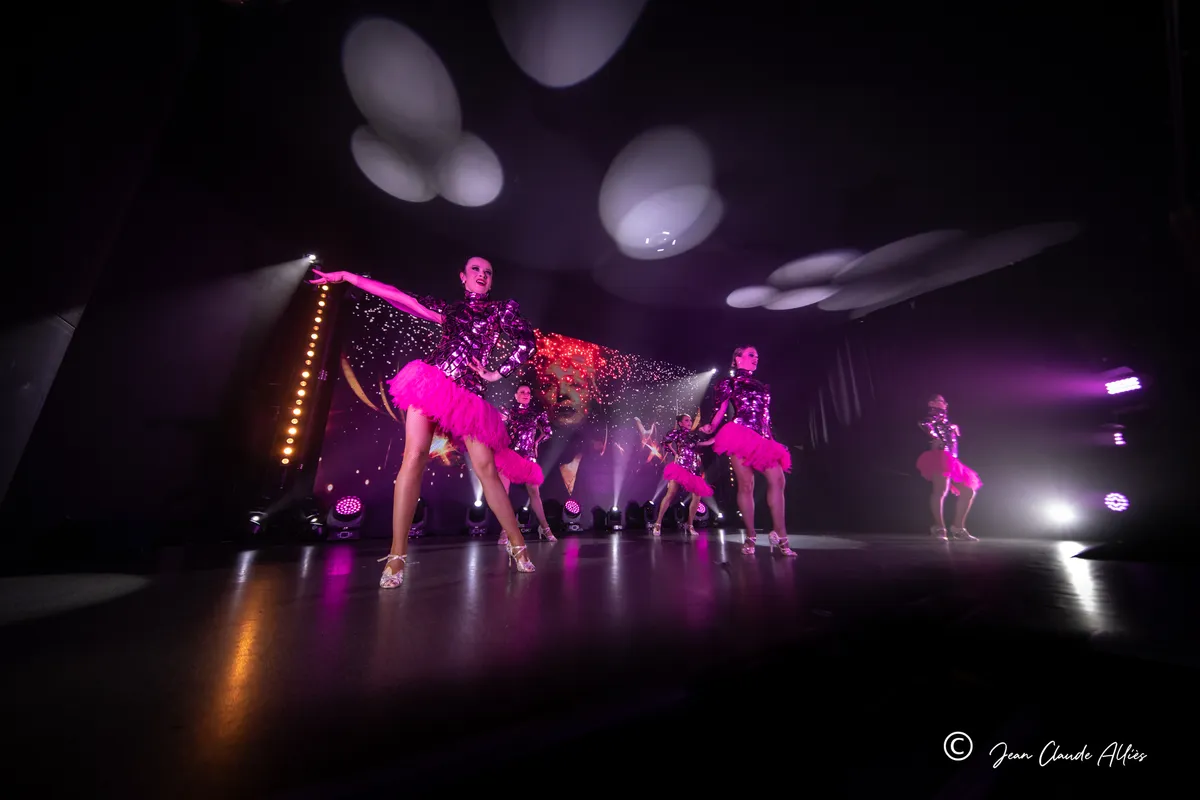 Danseuses en jupes roses et hauts brillants ouvrant les bras sur une scène éclairée en violet.