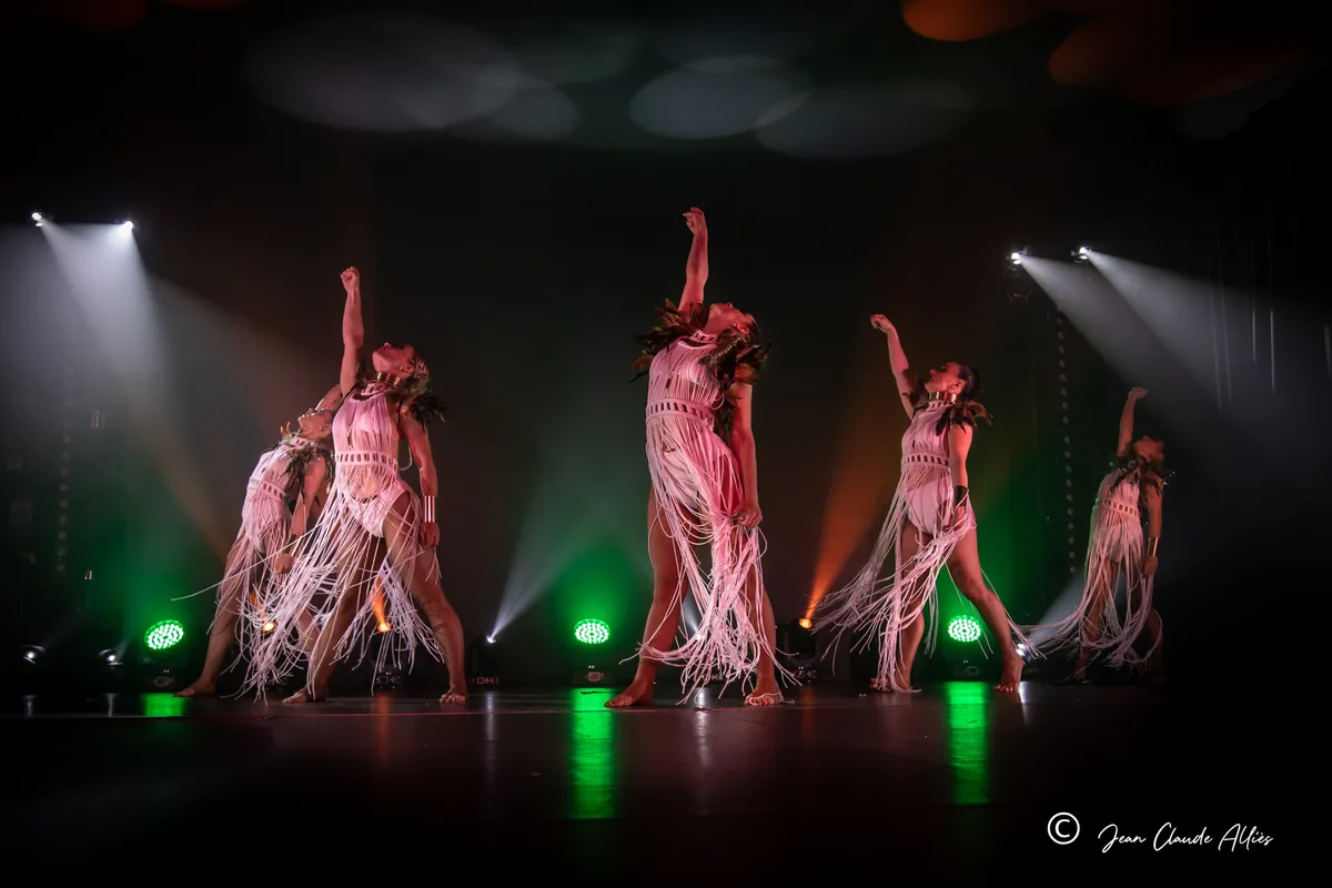 Danseuses en costumes à longues franges claires et coiffes de plumes, les bras levés sous une lumière verte.