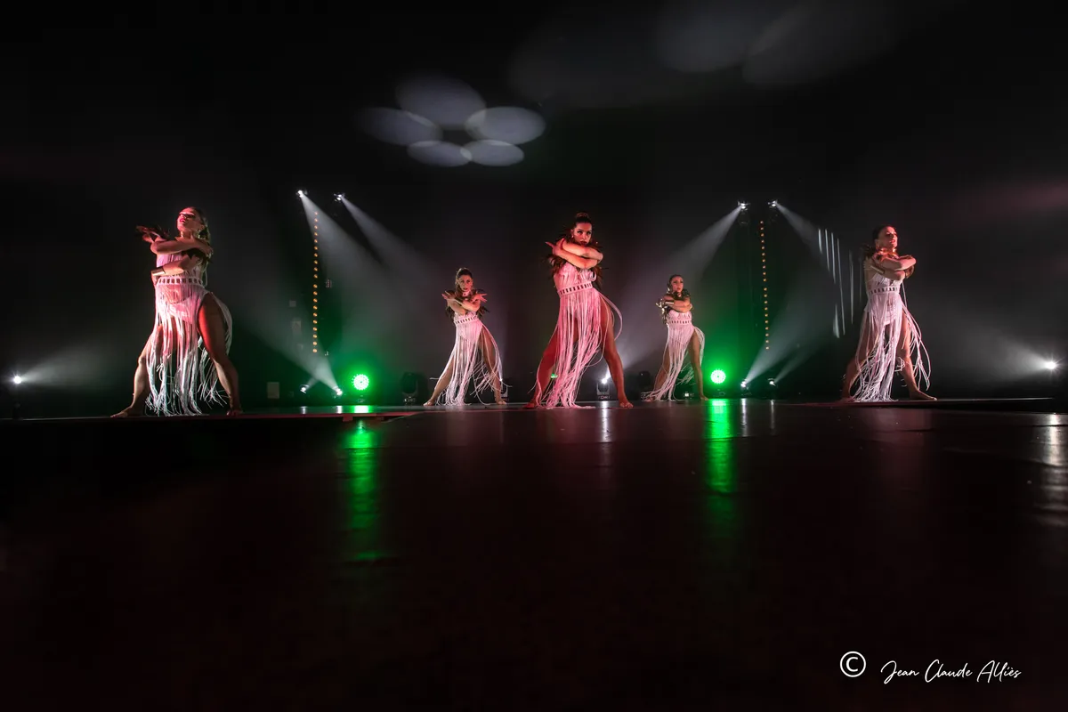 Vue large de danseuses en costumes à franges claires alignées sur scène entre des projecteurs verts et blancs.