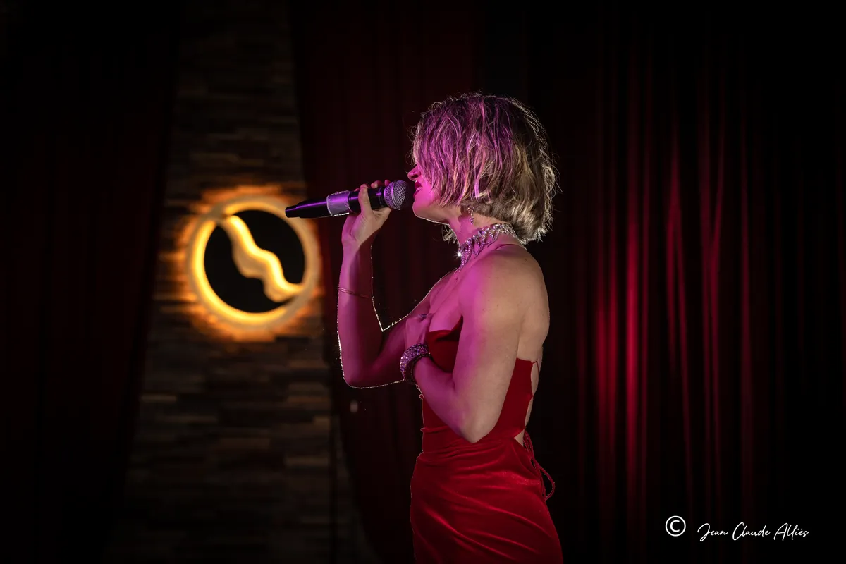Chanteuse aux cheveux courts en robe rouge, vue de profil au microphone près d'un rideau rouge.