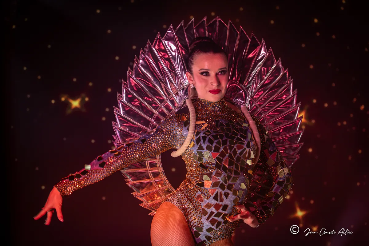 Danseuse en costume à facettes tendant un bras devant une parure argentée géométrique et un fond étoilé.