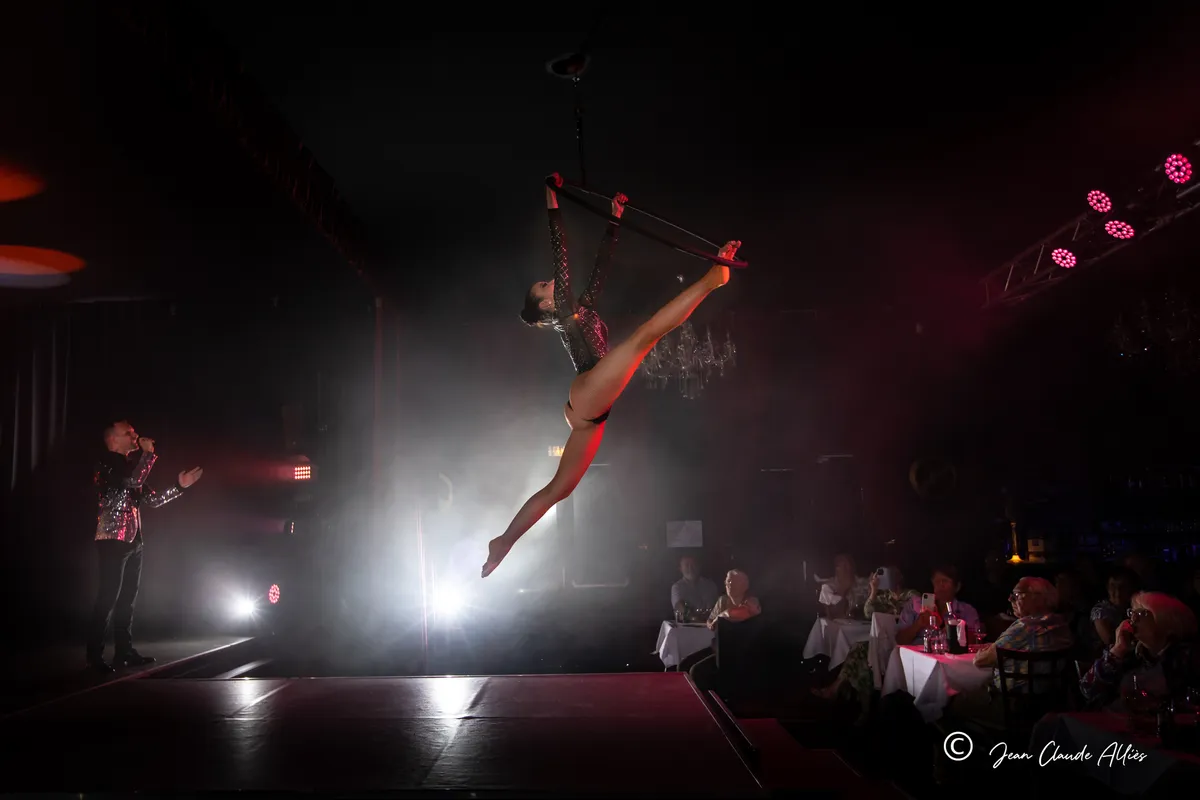 Acrobate suspendue à un cerceau aérien, une jambe tendue, au-dessus d'une scène entourée de tables.
