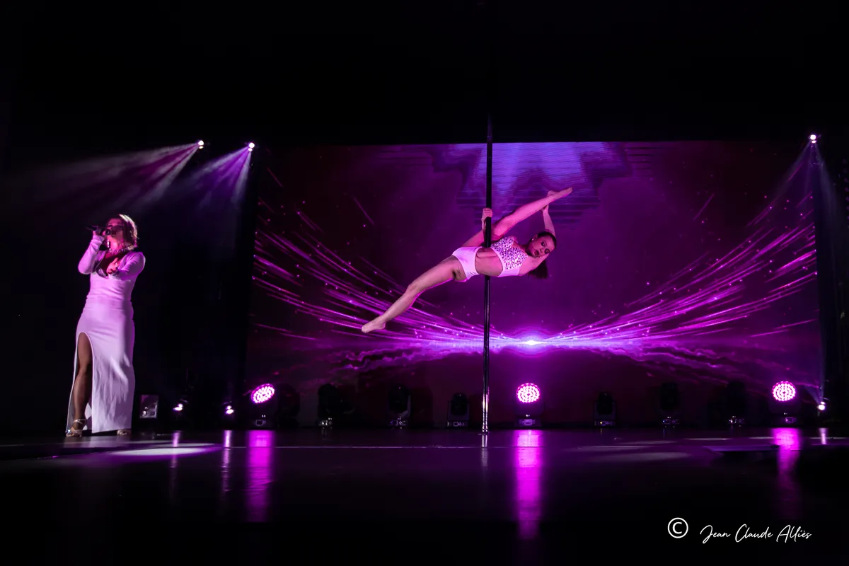 Acrobate en suspension sur une barre au centre de la scène, accompagnée d'une chanteuse en robe blanche.