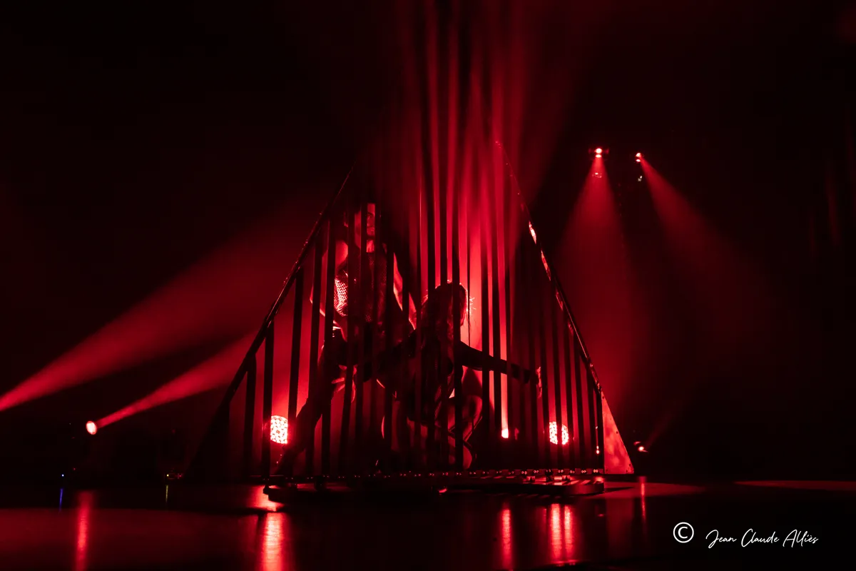Silhouettes de danseuses sur une structure à barreaux verticaux, éclairée par des faisceaux rouges.