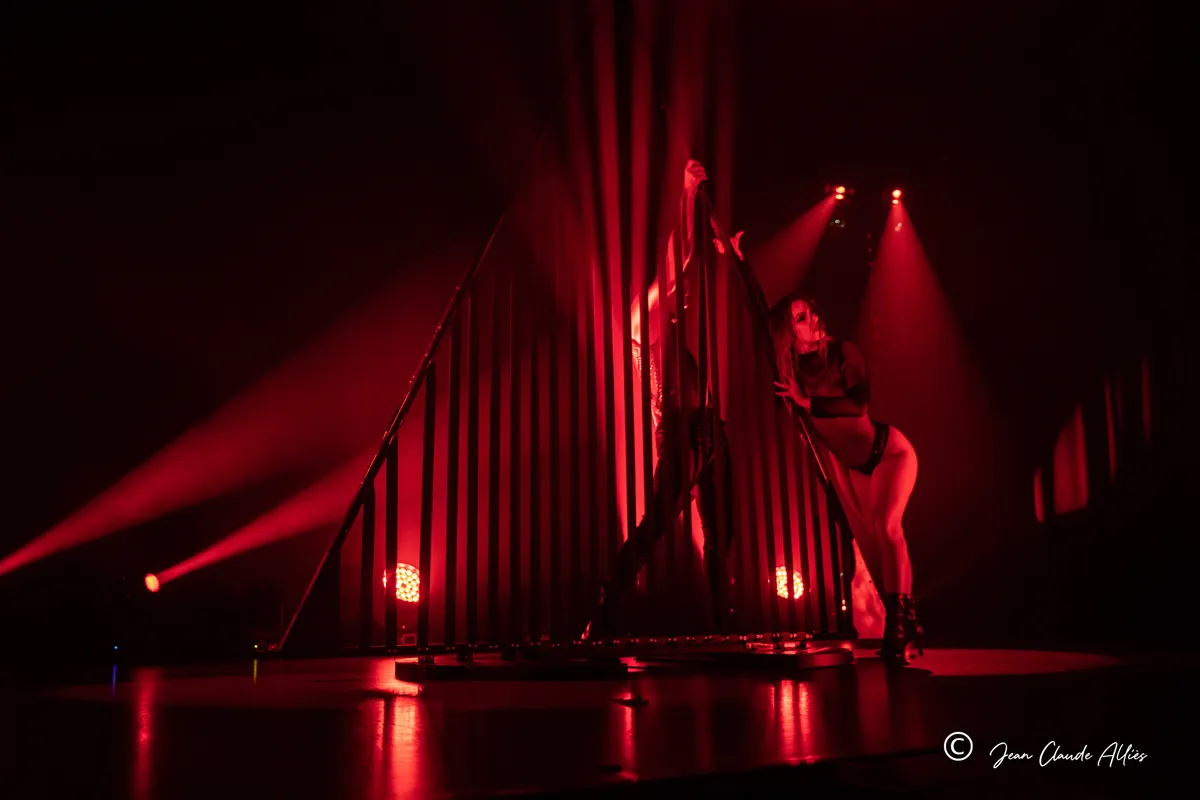 Danseuses réparties à plusieurs hauteurs sur une structure à barreaux, dans une lumière rouge intense.