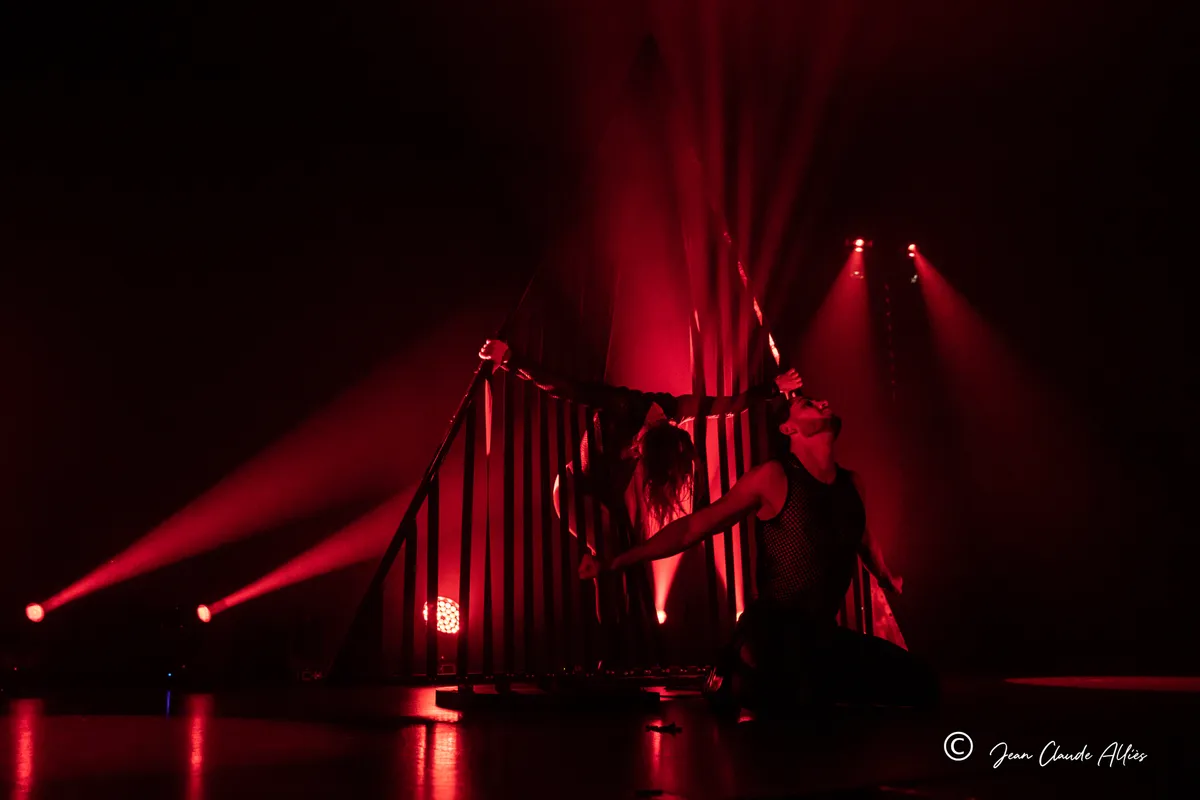 Danseuses étirées de part et d'autre d'une structure à barreaux, sous un éclairage rouge sur fond noir.