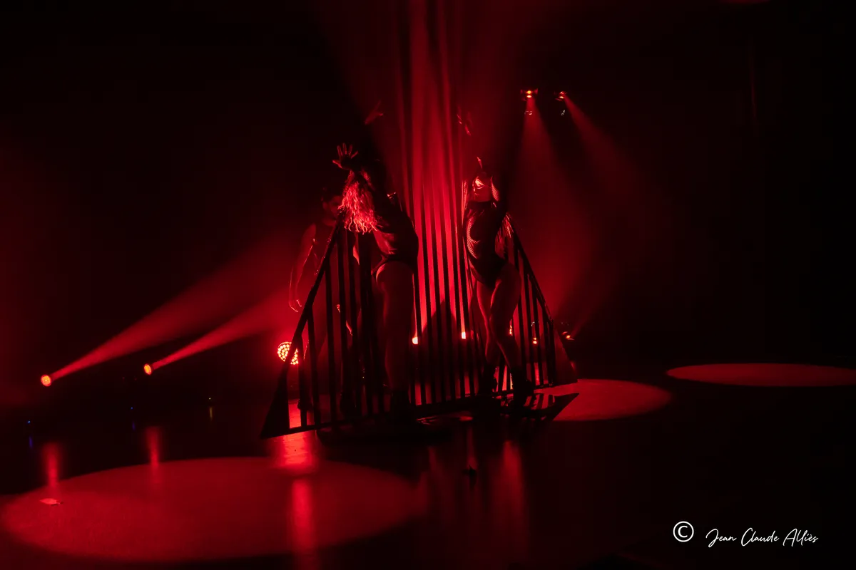 Vue large d'un tableau de danse autour d'une structure à barreaux, baignée de lumière rouge.