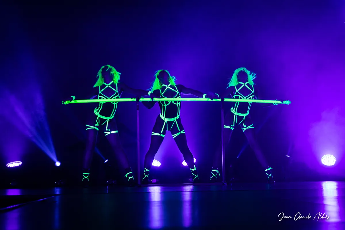 Trois danseuses au centre d'un tableau UV tenant une barre verte horizontale devant des projecteurs violets.
