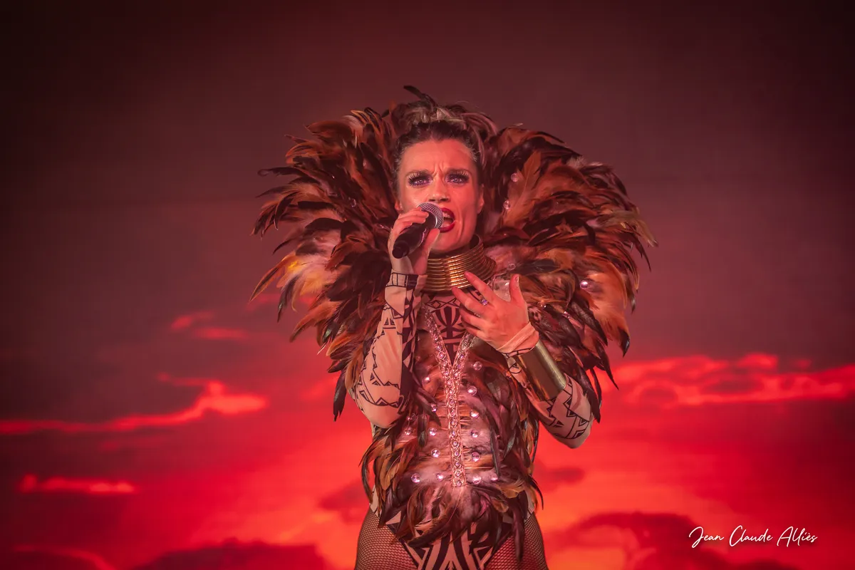 Chanteuse au micro encadrée d'une large parure de plumes brunes devant un fond rouge et orangé.